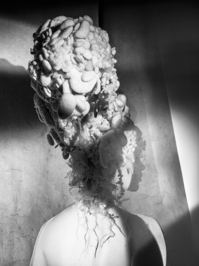 Monochrome profile of a person with an elaborate hairstyle or headdress, featuring bulbous, organic shapes in soft, diffused lighting.
