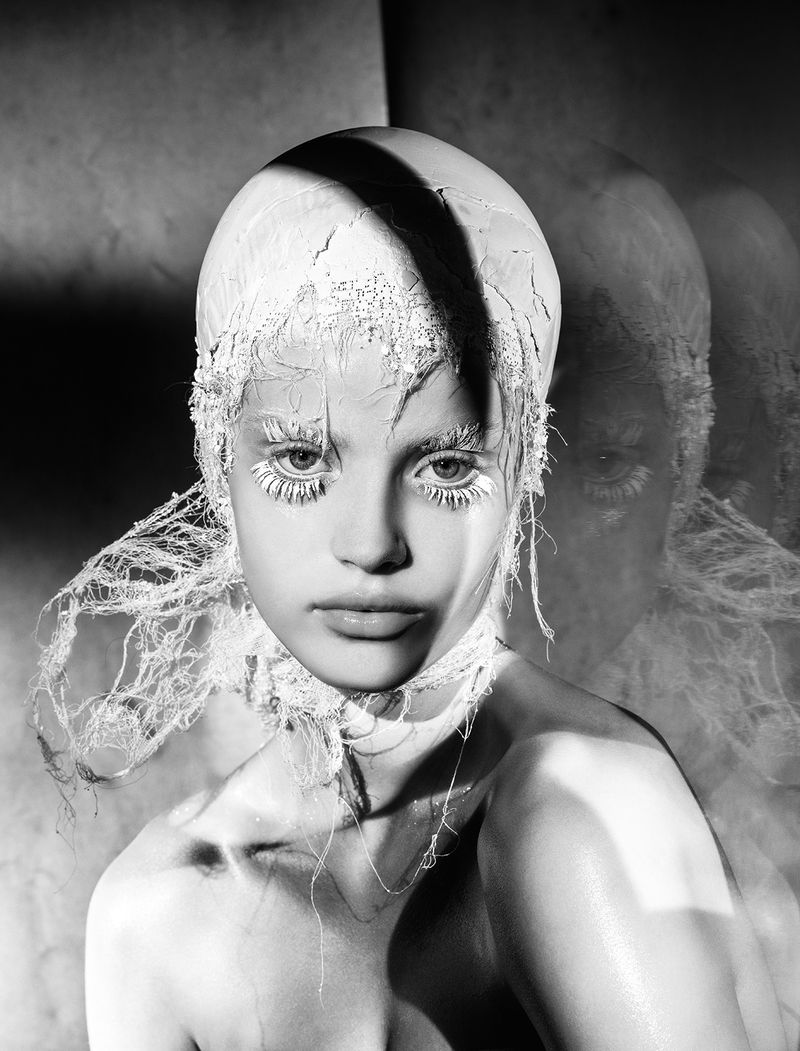 Monochrome portrait of a young woman with elaborate makeup, head covering, and white eyelashes, set against a dark, moody background.