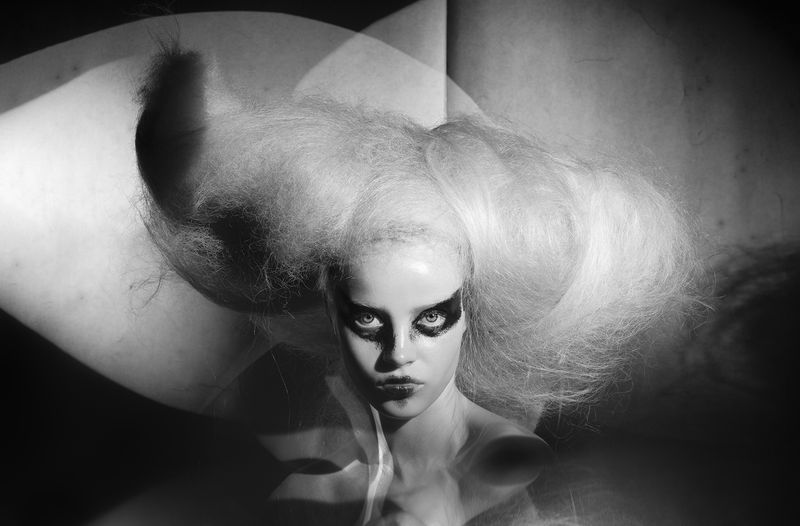 Striking black and white portrait of a woman with dramatic makeup and voluminous hairstyle against a textured, abstract background.