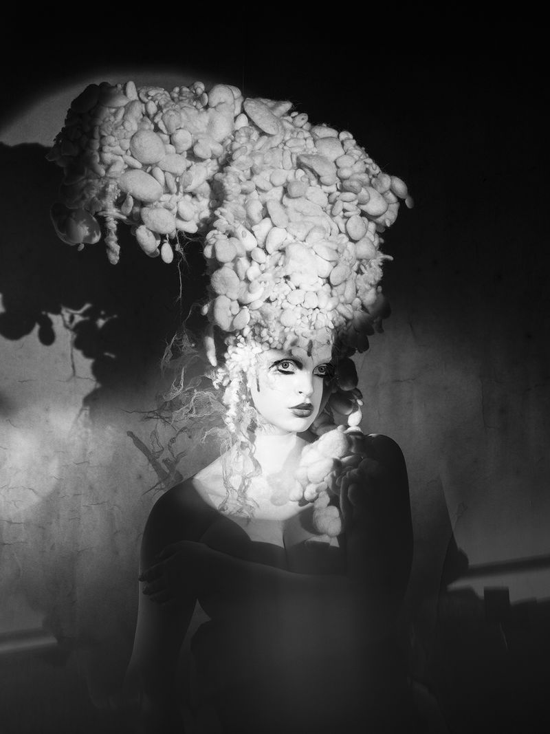 Artistic black and white portrait of a person with dramatic makeup and elaborate headwear standing in a dimly lit, textured environment.