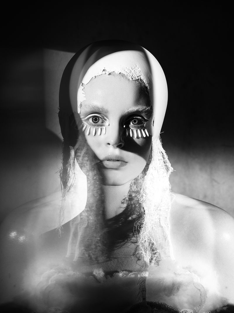 Striking monochrome portrait of a woman with dramatic eye makeup, styled with a white headpiece, set against a shadowy backdrop.