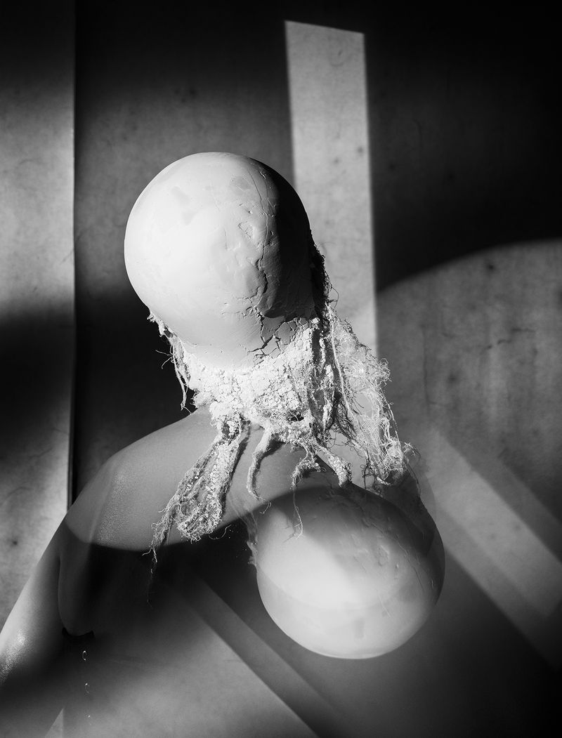 Monochrome abstract artwork featuring a bulbous, textured form with draped fabric, set against a starkly lit background with geometric shadows.
