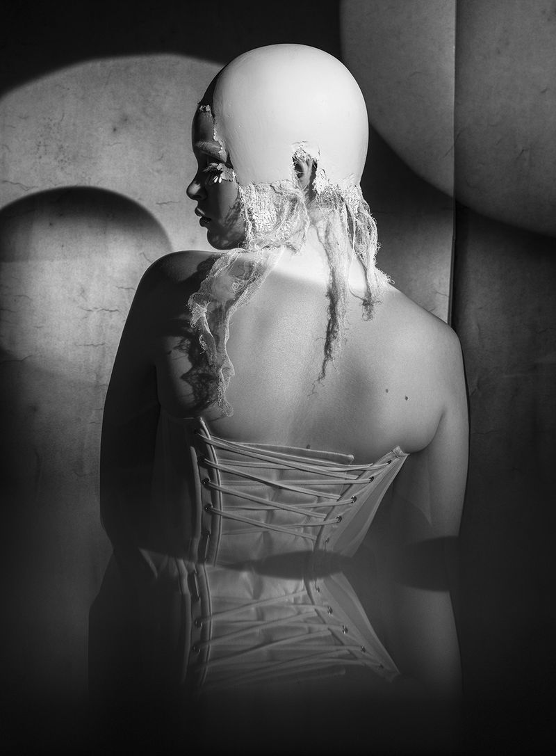 Monochrome portrait of a person wearing a white head covering and corset, standing in a dimly lit room with textured walls.