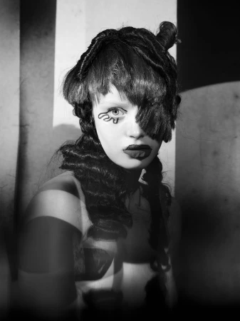 Monochrome portrait of a young woman with dark, stylized hair, dramatic eye makeup, and lipstick in a studio setting with striped shadows.