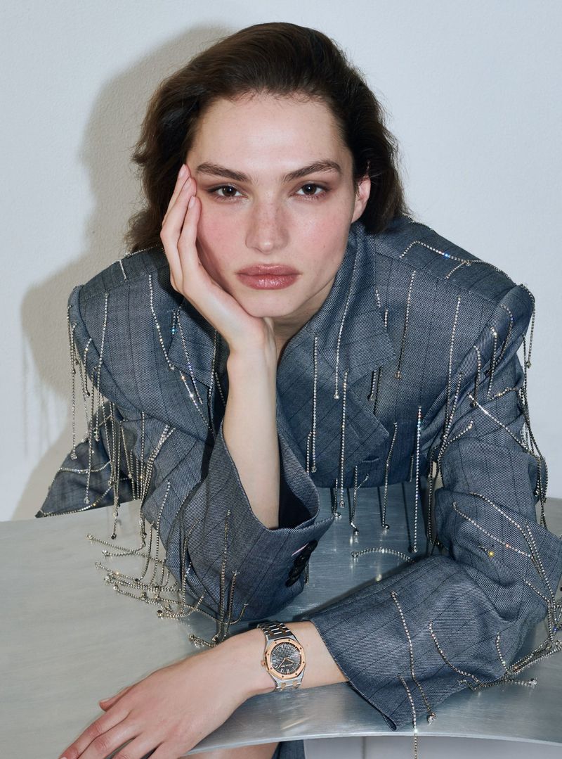 Portrait of a woman wearing a grey embellished blazer with an elegant watch, posing in a minimalist indoor studio environment.