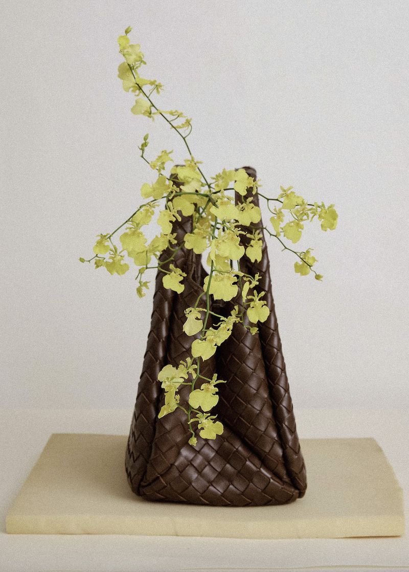 Brown woven leather tote bag with delicate yellow orchids arranged inside, creating an elegant fashion and floral display against a white background.