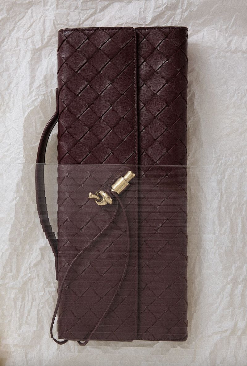 Close-up of a woven leather wallet in a dark purple color, resting on a textured off-white surface with soft lighting enhancing the details.