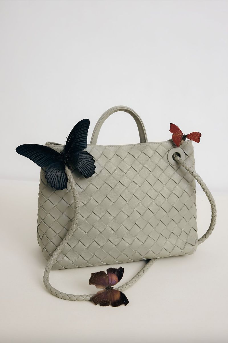 Light grey woven leather handbag with braided strap and butterfly embellishments in a studio setting against a white backdrop.