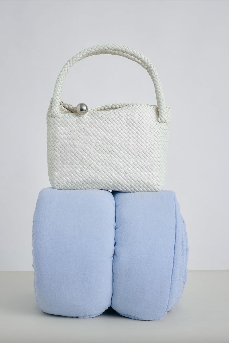 Ivory woven handbag with a round top handle and silver clasp placed atop light blue cylindrical cushions, studio shot.