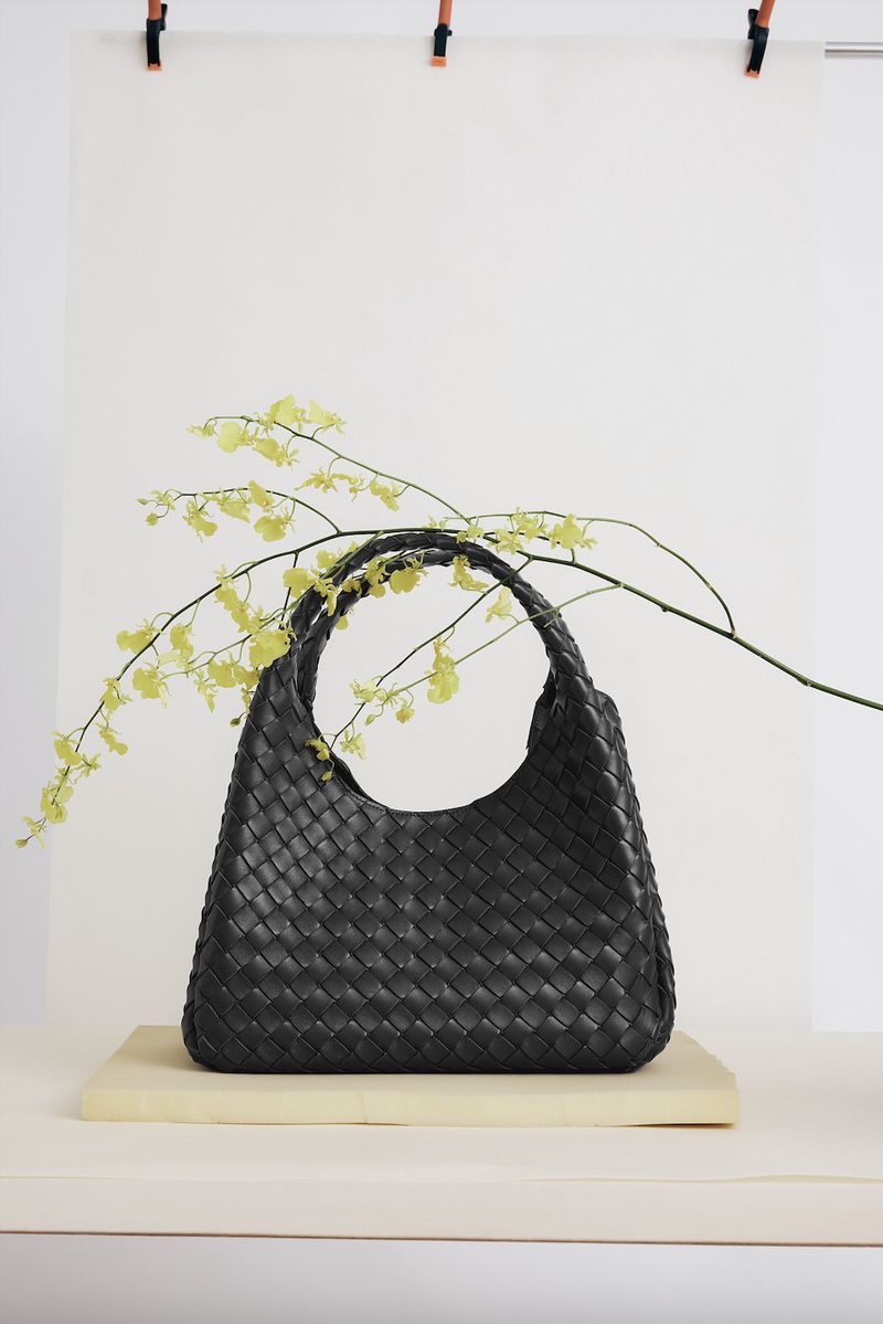 Woven black handbag on a light yellow surface with a sprig of delicate green flowers set against a bright white backdrop.