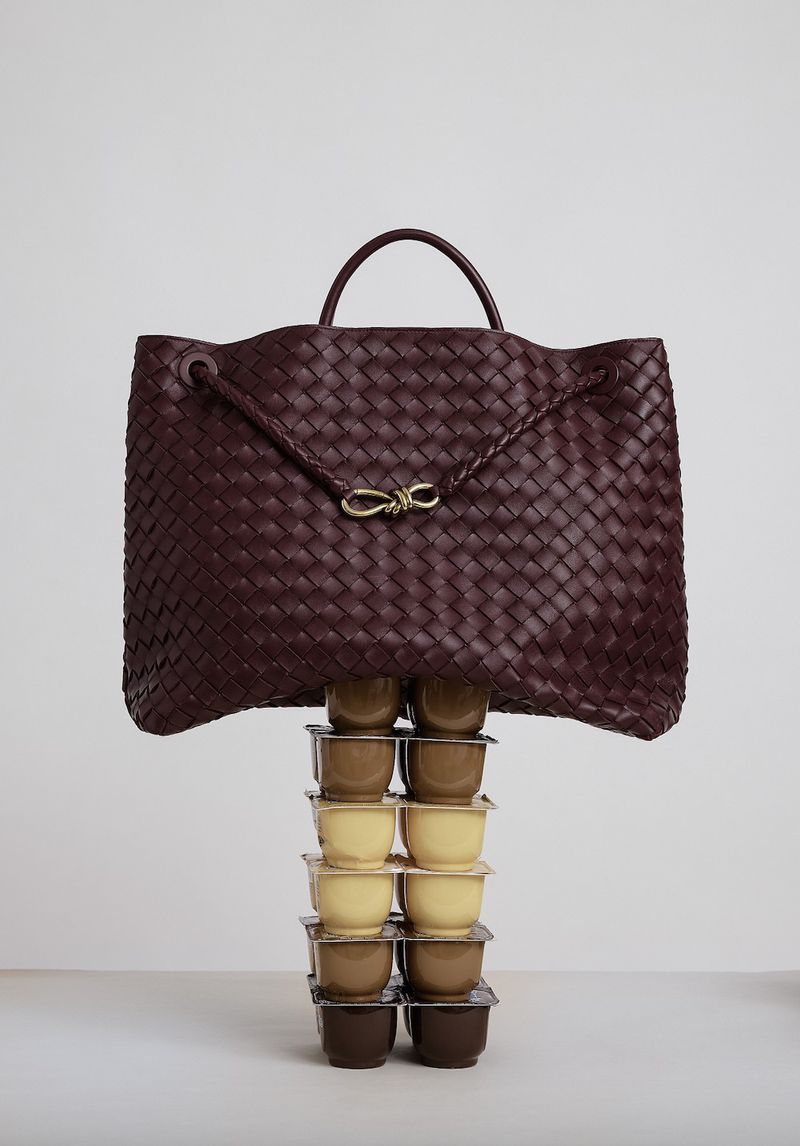 Burgundy woven leather handbag on a pedestal of stacked dessert cups, set against a clean, neutral backdrop in a still life product shot.