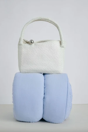 White woven top-handle handbag with a silver spherical clasp resting on a light blue padded cushion in a studio setting.