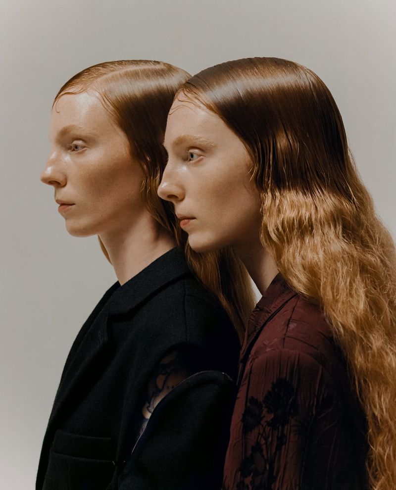 Two women with slicked-back red hair stand side by side in a portrait. One wears a black jacket, and the other wears a dark red shirt.