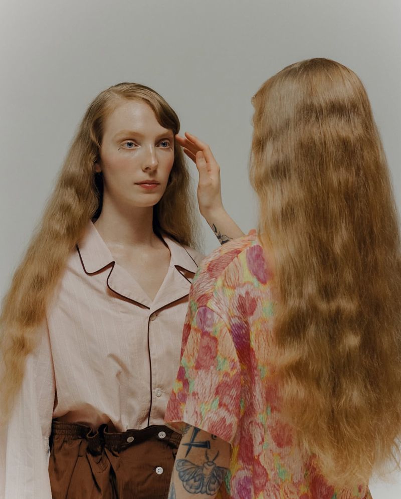 A model in a pale pink collared shirt touched on the temple by a woman with long wavy hair and arm tattoos against a neutral backdrop.