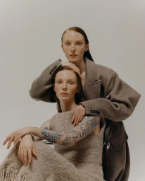 Two female models posing together in neutral knitwear and an oversized wool coat against a plain studio backdrop.