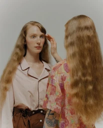 A model in a pale pink collared shirt touched on the temple by a woman with long wavy hair and arm tattoos against a neutral backdrop.
