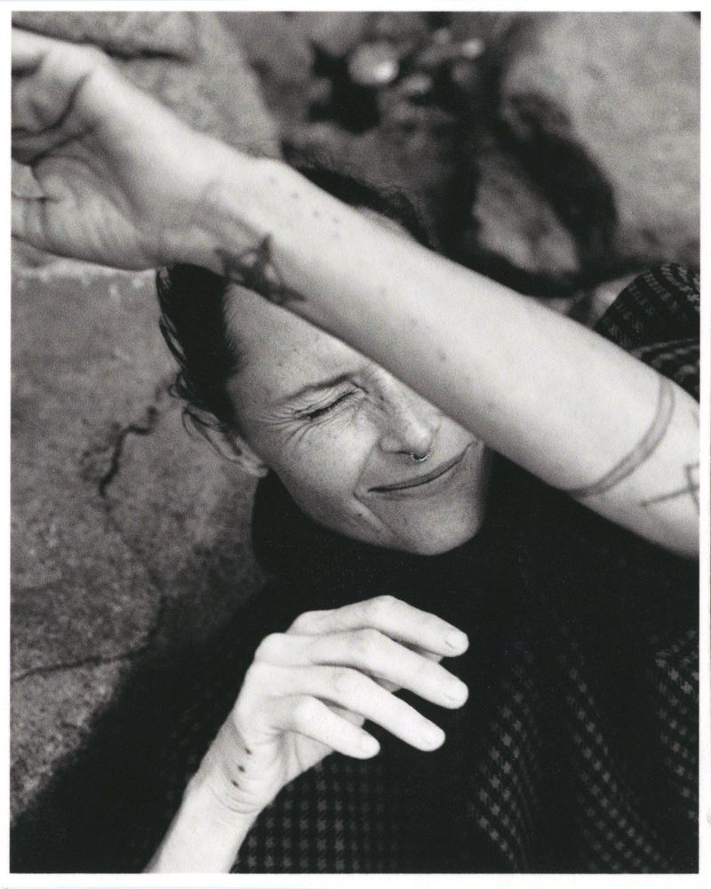 Monochrome portrait of an adult woman with a nose ring and arm tattoos, covering her eyes with her arm in an outdoor setting.