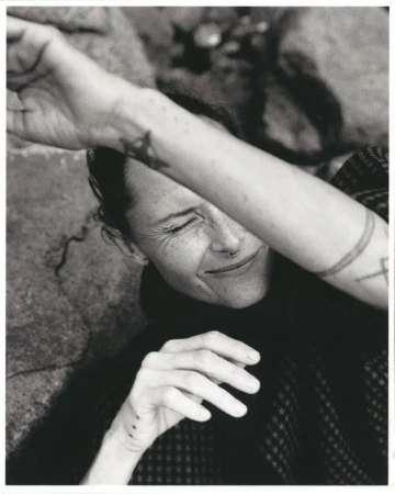 Monochrome portrait of an adult woman with a nose ring and arm tattoos, covering her eyes with her arm in an outdoor setting.
