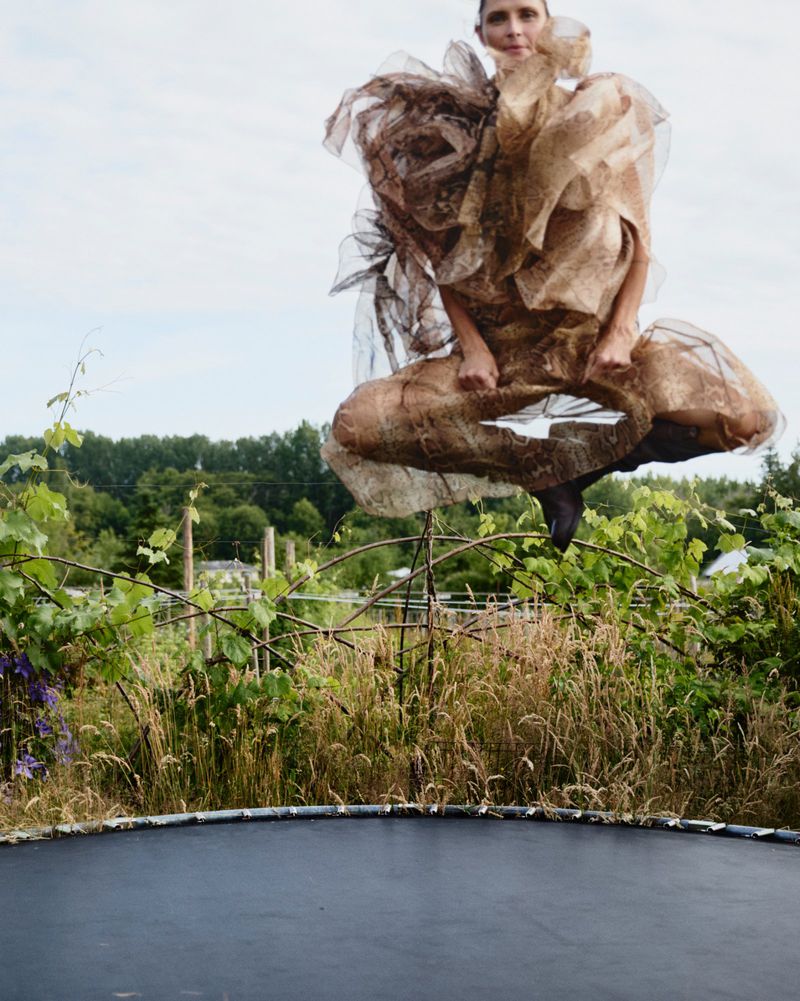 A woman in an elaborate tan ruffled dress leaps on a trampoline surrounded by lush green vegetation and a bright blue sky in an outdoor setting.