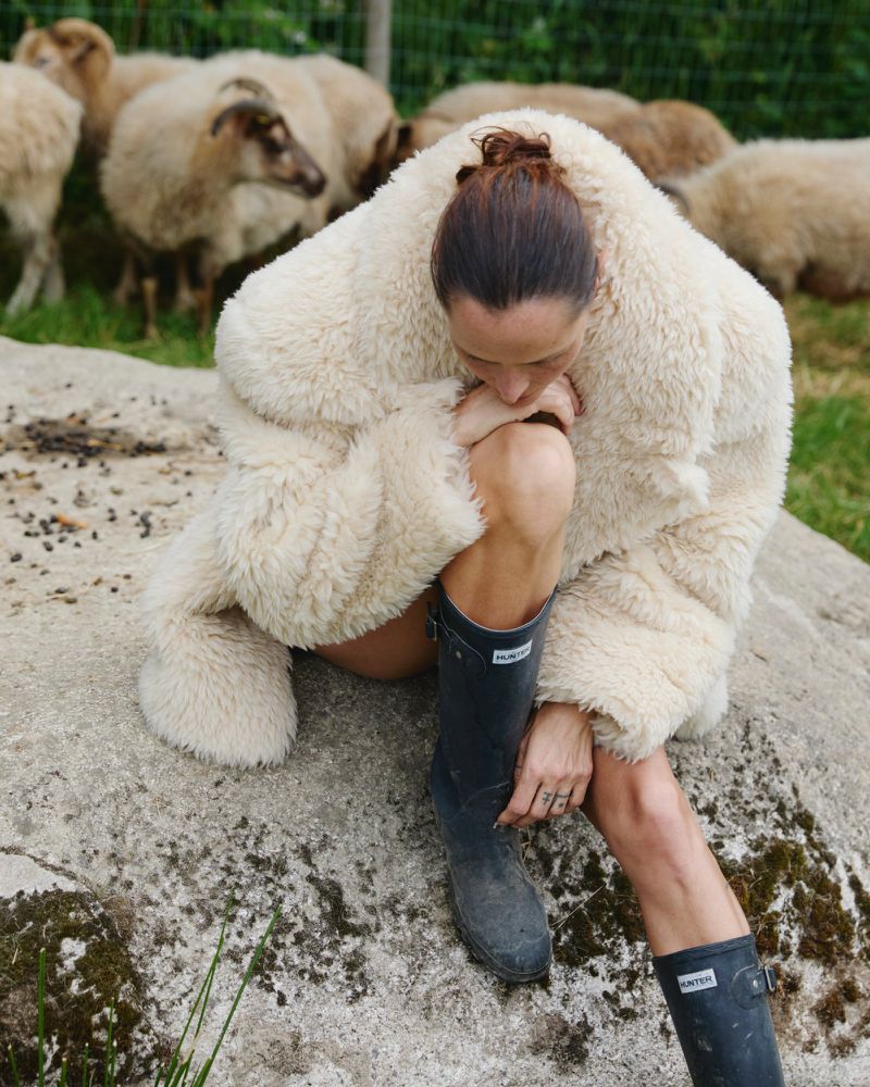 Woman in a sheepskin jacket and boots sits on a rock in a grassy field with sheep grazing in the background. Outdoor setting, overcast light.