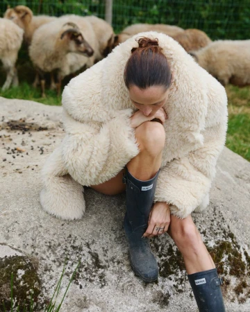 Woman in a sheepskin jacket and boots sits on a rock in a grassy field with sheep grazing in the background. Outdoor setting, overcast light.
