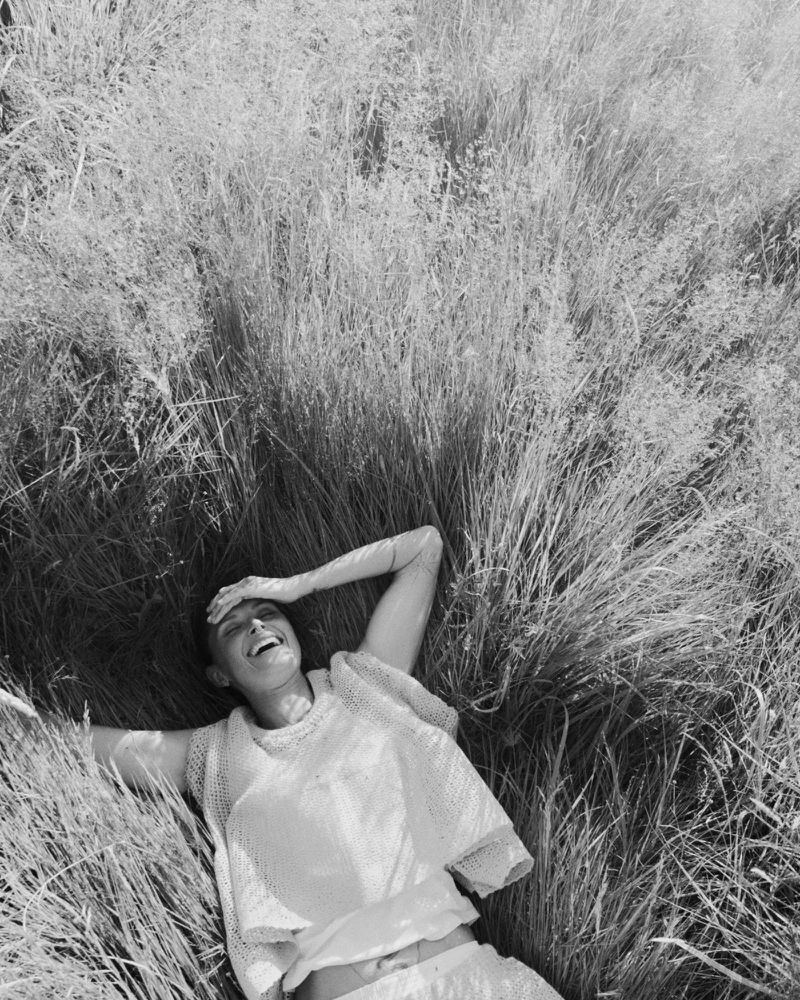 Monochrome photo of a smiling woman lying in tall grass, shielding her eyes, in soft, diffused lighting.