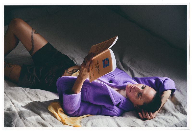 Woman in purple coat reads a book while laying on a bed with a yellow scarf, creating a cozy and intimate scene.