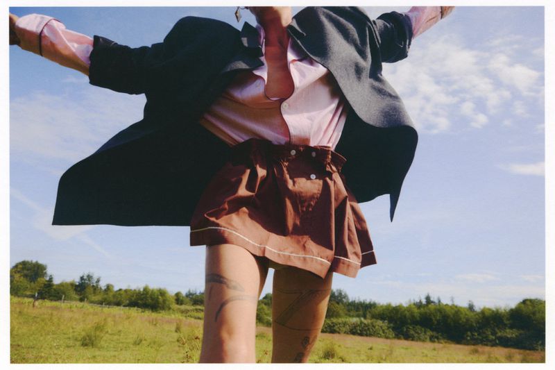 Model Tasha Tilberg standing in an open grassy meadow with arms raised, wearing an oversized dark jacket, pink shirt, and brown skirt.