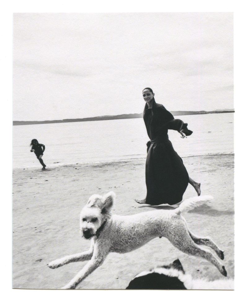 Tasha Tilberg walking barefoot on a sandy beach in a dark coat as a white dog bounds past in the foreground.