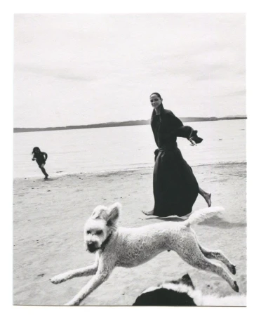 Tasha Tilberg walking barefoot on a sandy beach in a dark coat as a white dog bounds past in the foreground.