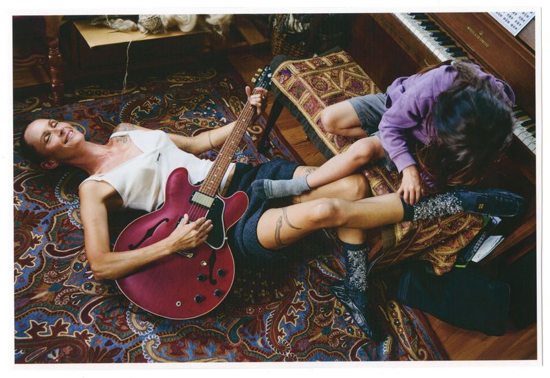 Adult woman plays red electric guitar lying on a patterned rug indoors, while a child in a purple shirt sits nearby, smiling in daylight.