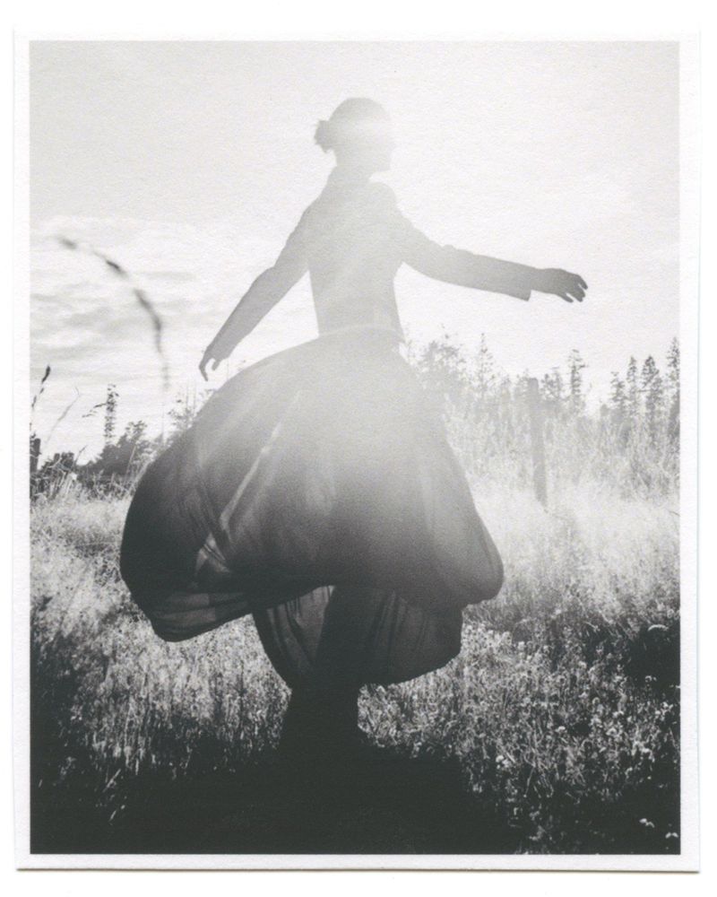 Monochromatic portrait of a person with arms outstretched, wearing a vintage dress, standing in a field with trees in the background.