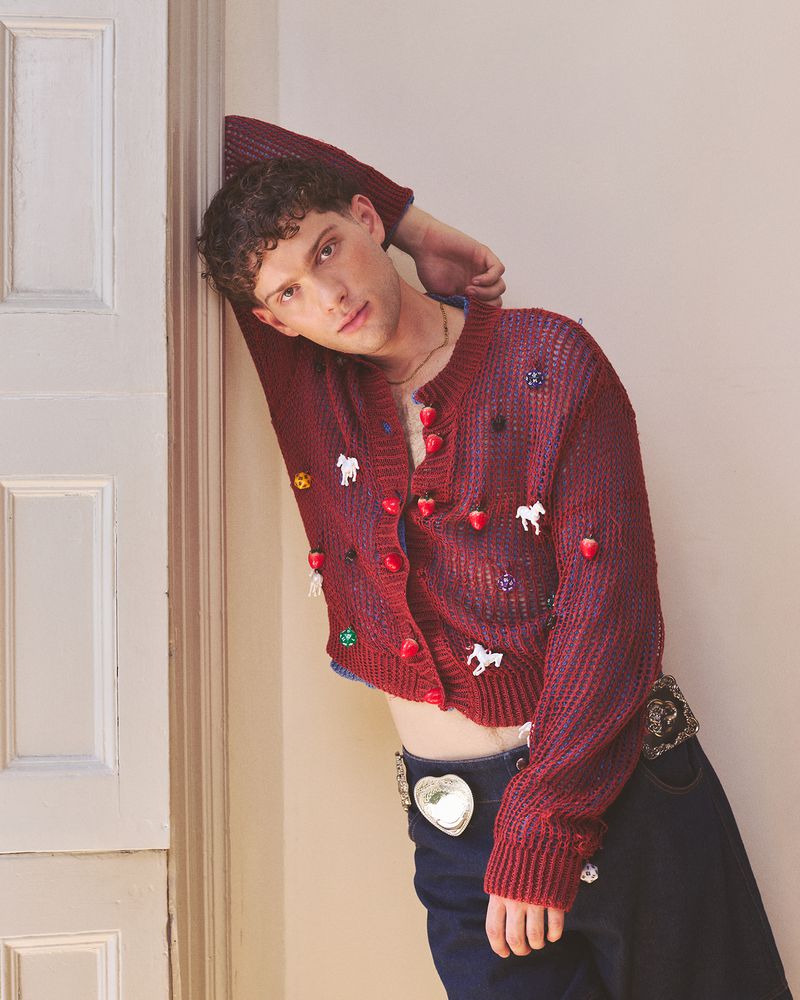 Young adult man wearing a novelty cardigan sweater with strawberry and horse embellishments, leans against a white wall in a studio.