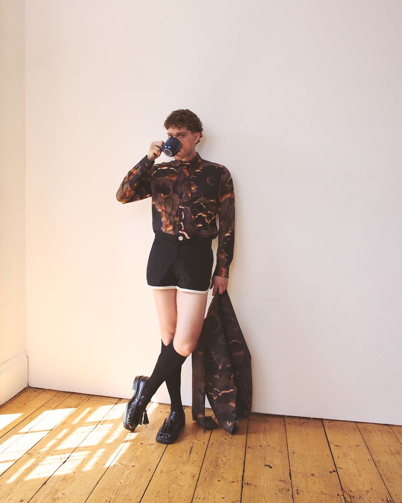Young adult with curly hair wears patterned shirt and shorts while drinking from a mug inside a room with wood floors.
