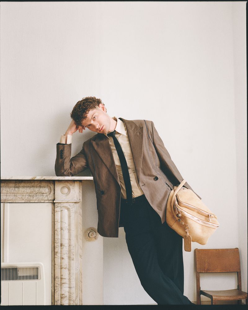 Leo Reich wearing a brown oversized blazer and black trousers, leaning against a vintage fireplace holding a tan leather bag.