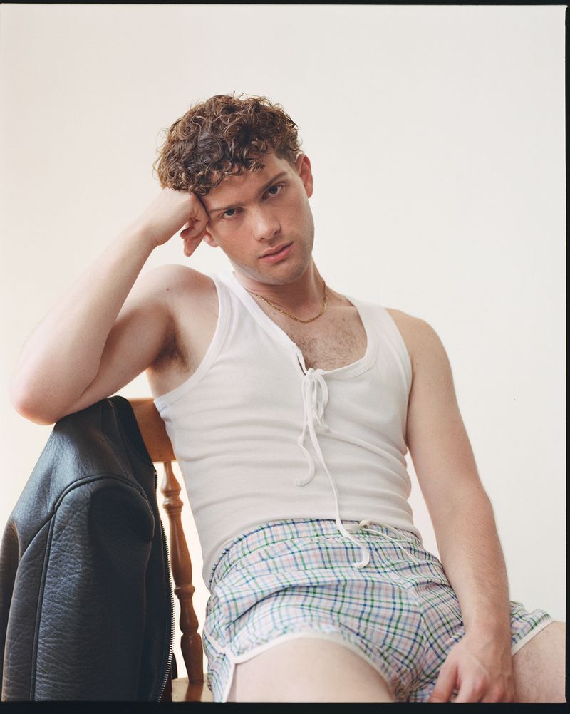 Leo Reich seated on a wooden chair wearing a white lace-up tank top and plaid shorts with a black leather jacket hanging nearby.