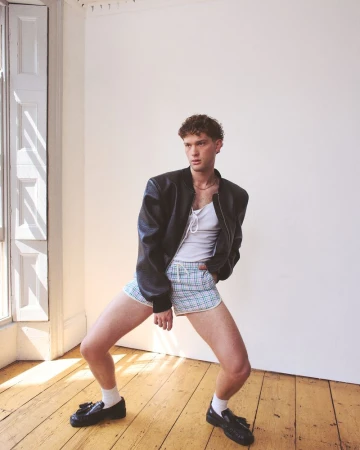Young adult male in an unbuttoned leather jacket and shorts poses in a brightly lit indoor setting near a window, gazing off to the side.