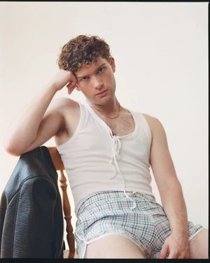 Leo Reich seated on a wooden chair wearing a white lace-up tank top and plaid shorts with a black leather jacket hanging nearby.