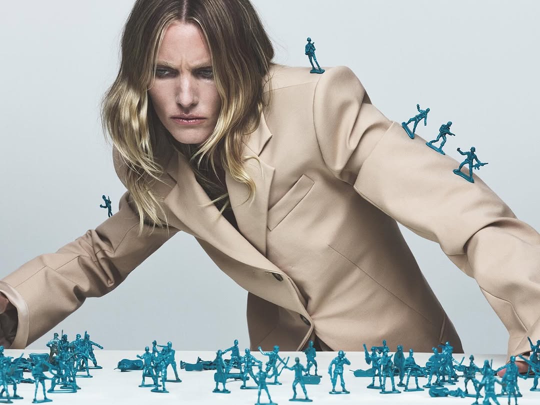 Model in a beige blazer surrounded by small, blue toy soldiers on a white surface in a studio setting under bright lighting.