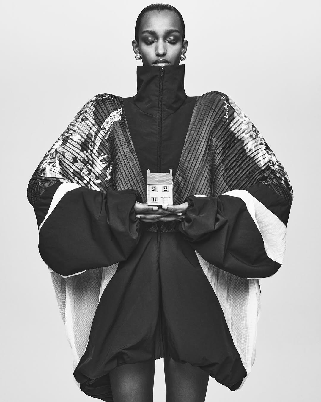 Fashion shot of a model wearing voluminous black clothing and holding a miniature house in a monochromatic color scheme against light backdrop.
