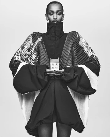 Fashion shot of a model wearing voluminous black clothing and holding a miniature house in a monochromatic color scheme against light backdrop.