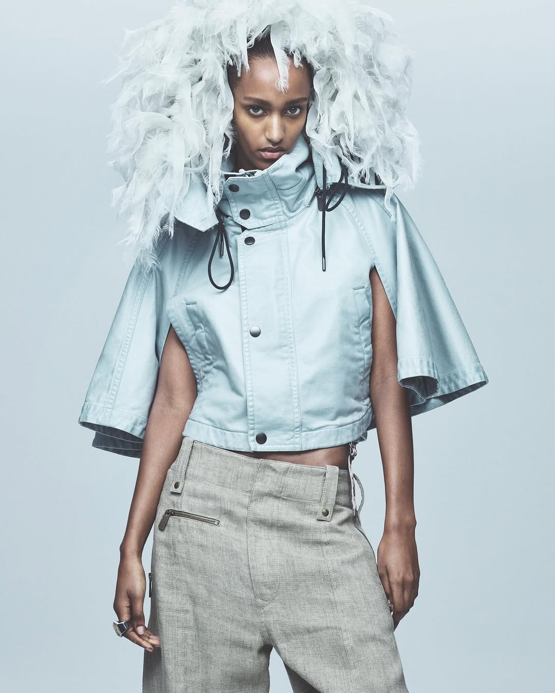 Fashion portrait of a Black woman with a feathered headpiece and blue jacket, posing in a photo studio with neutral background and lighting.