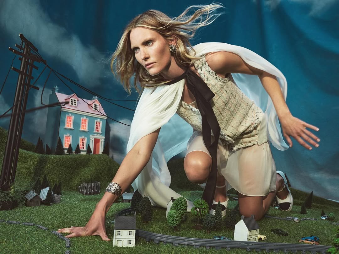 Fashion editorial shot of a blonde adult woman in a light-colored dress and cape kneeling on a miniature landscape with small houses.