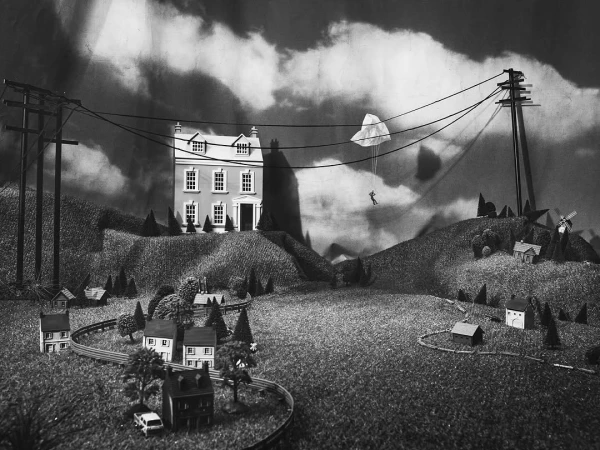 Monochrome diorama featuring a house on a hill, power lines, parachutist, and toy town, evoking a surreal and miniature world.