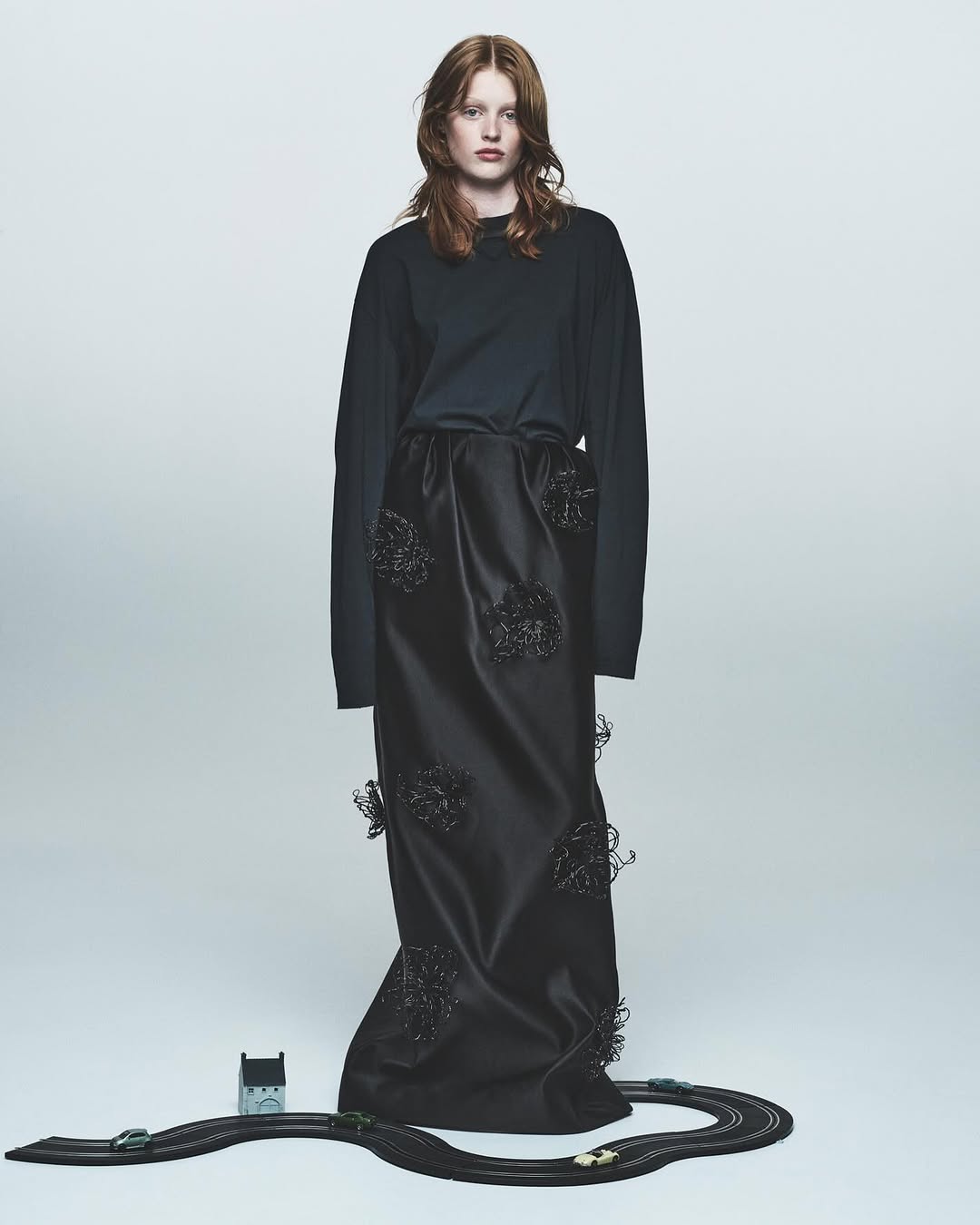 Fashion shot of a red-haired woman in a long-sleeved black shirt and long black skirt with toy race track at her feet.