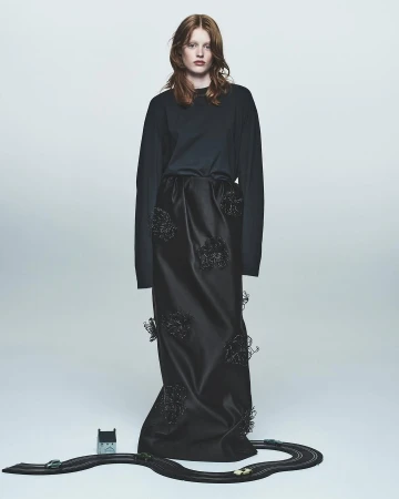Fashion shot of a red-haired woman in a long-sleeved black shirt and long black skirt with toy race track at her feet.
