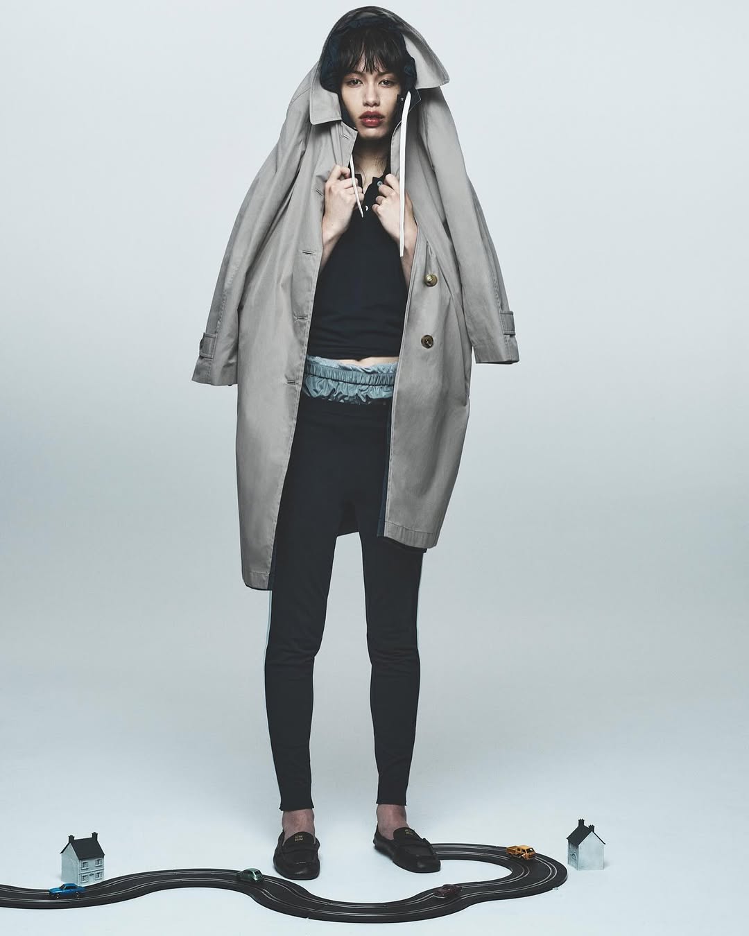 Editorial fashion shot of a stylish woman posing indoors with a trench coat, black top, leggings, and toy race track.