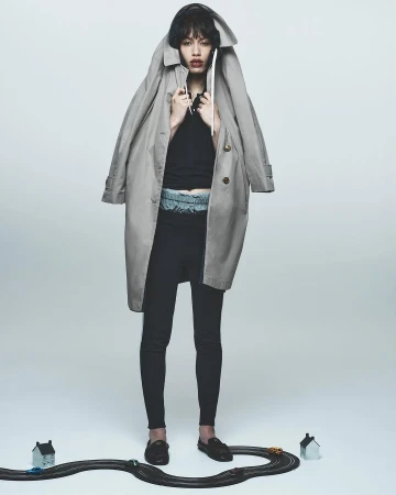 Editorial fashion shot of a stylish woman posing indoors with a trench coat, black top, leggings, and toy race track.
