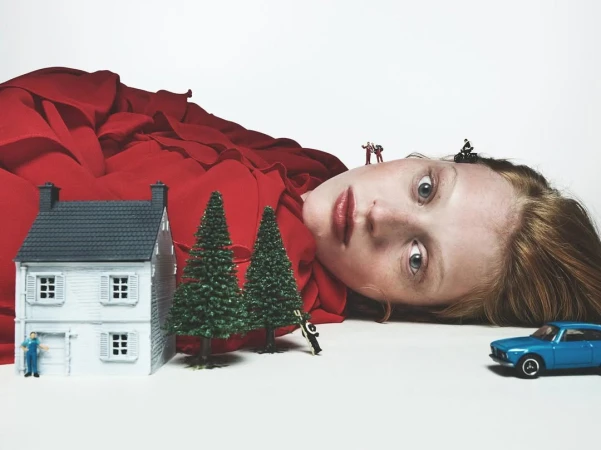 Surreal composition featuring a red-dressed woman's face, toy house, miniature people, and blue car on a white background.