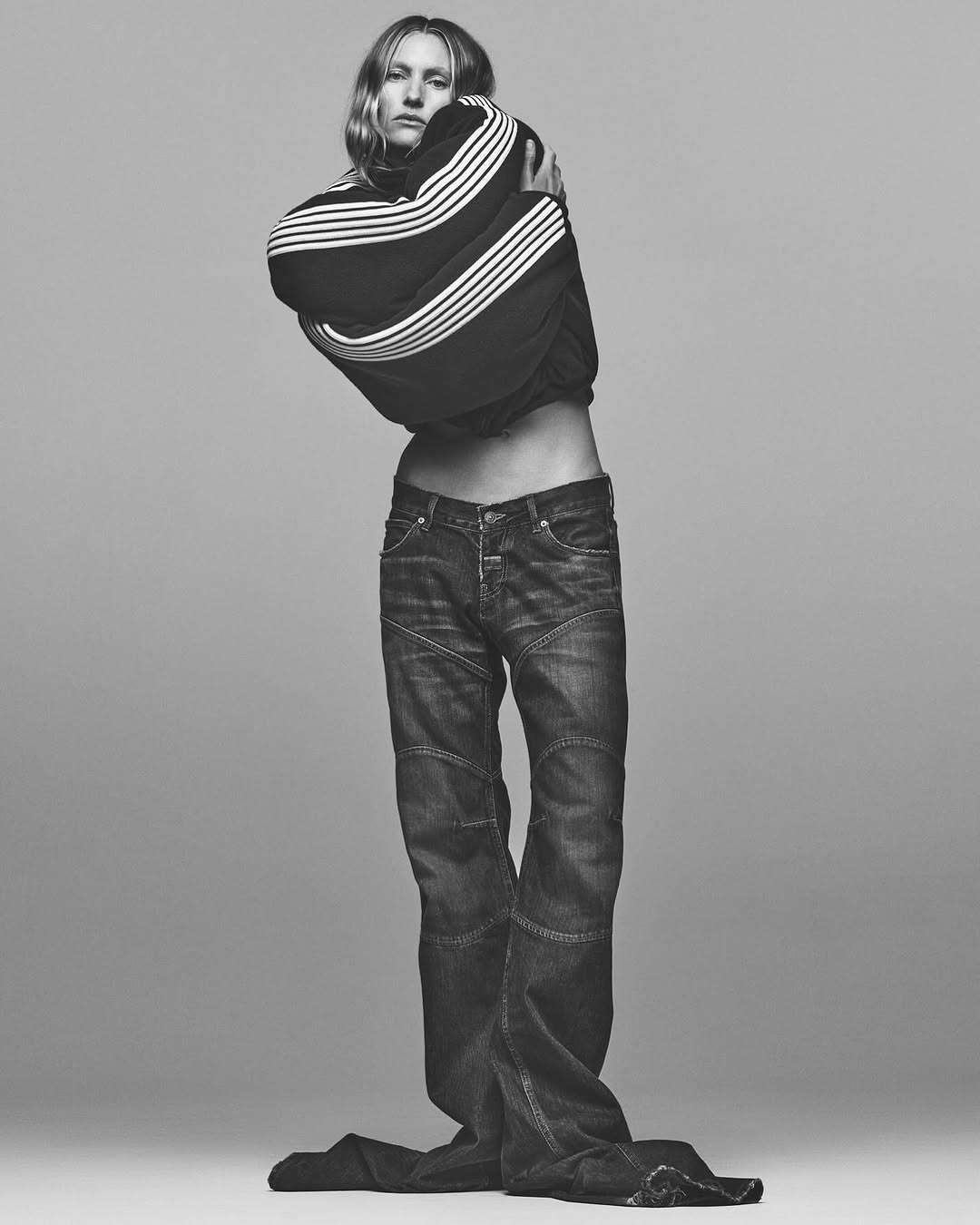Full body studio shot of a person in dark jeans and a striped sweatshirt. The studio background is a neutral grey tone.